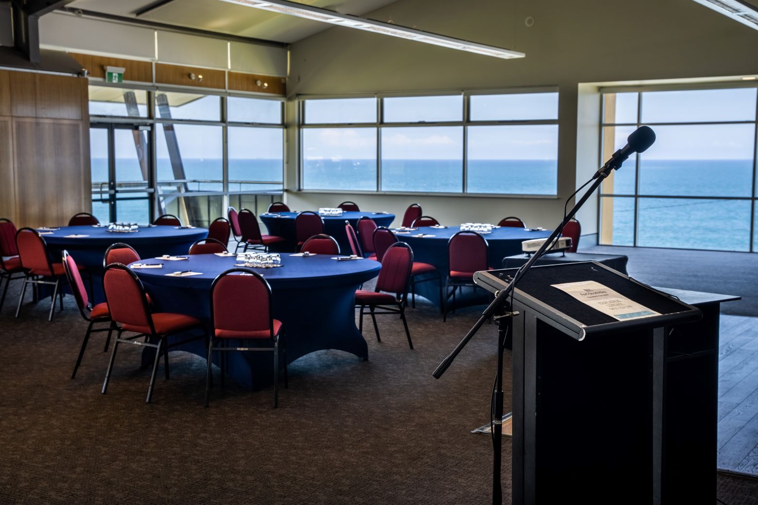 Fort Scratchley Function Centre set up cabaret style for a business event, with round tables and chairs arranged to face a microphone and lectern at the front of the room, and large windows showing the ocean view at the back of the room.