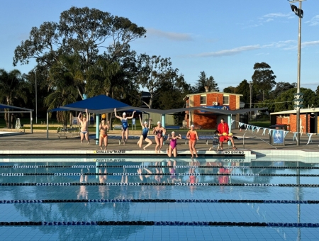 A group of swimmers in a line, mid-air as they jump into the pool.