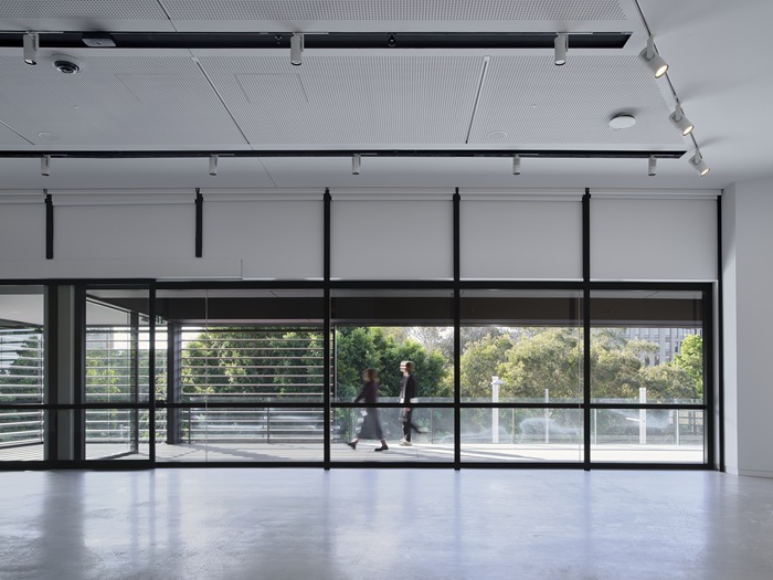 Photograph showing empty exhibition space, looking through glass windows to see two people walking outside in the daylight